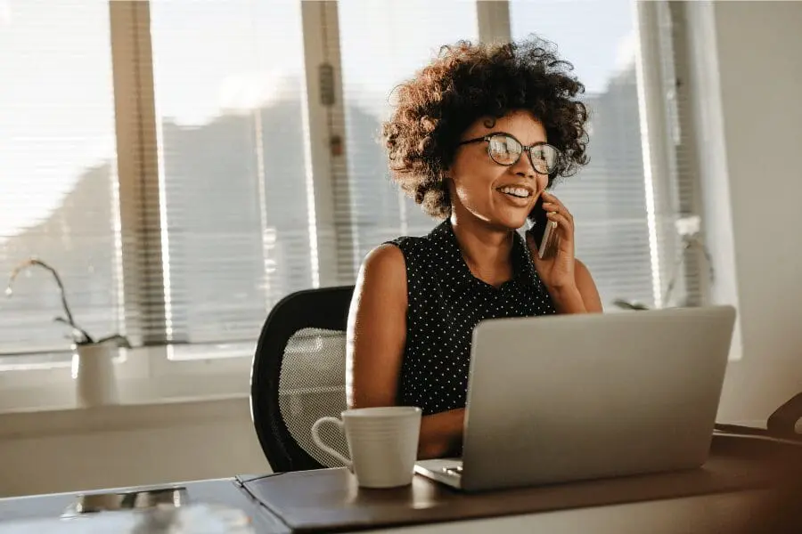 Une femme souriante aux cheveux bouclés et aux lunettes est assise à un bureau avec un ordinateur portable et parle au téléphone. La lumière du soleil traverse les stores derrière elle et une tasse de café est posée sur le bureau.