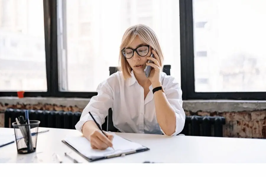 Une femme portant des lunettes et une chemise blanche est assise à un bureau, parlant au téléphone tout en écrivant dans un carnet. Elle se trouve dans un bureau lumineux avec de grandes fenêtres derrière elle.