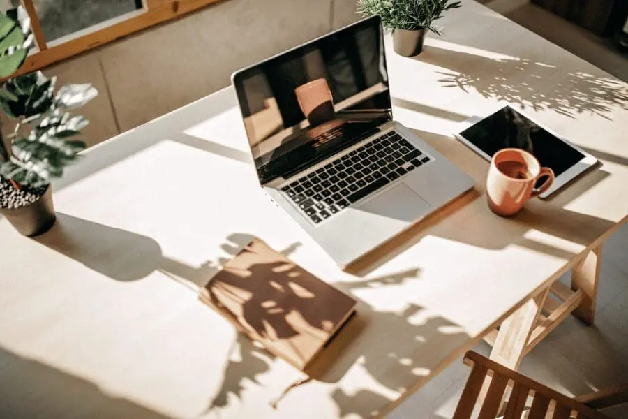Un bureau ensoleillé avec un ordinateur portable, une tablette, un carnet de notes, une tasse à café et des plantes en pot. La lumière du soleil et les ombres des plantes créent une atmosphère chaleureuse et accueillante dans l'espace de travail.