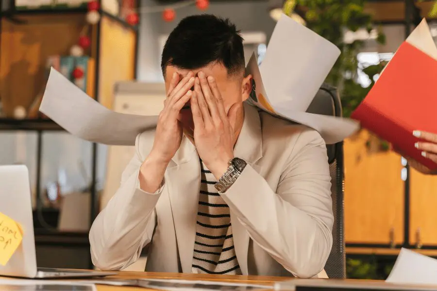 Un homme vêtu d'un blazer beige est assis à un bureau, le visage entre les mains, l'air stressé. Des papiers volent autour de lui, suggérant un environnement de travail agité ou surchargé.