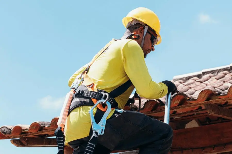 Un ouvrier du bâtiment portant un casque jaune et un harnais de sécurité grimpe sur une échelle pour travailler sur un toit en tuiles sous un ciel bleu.