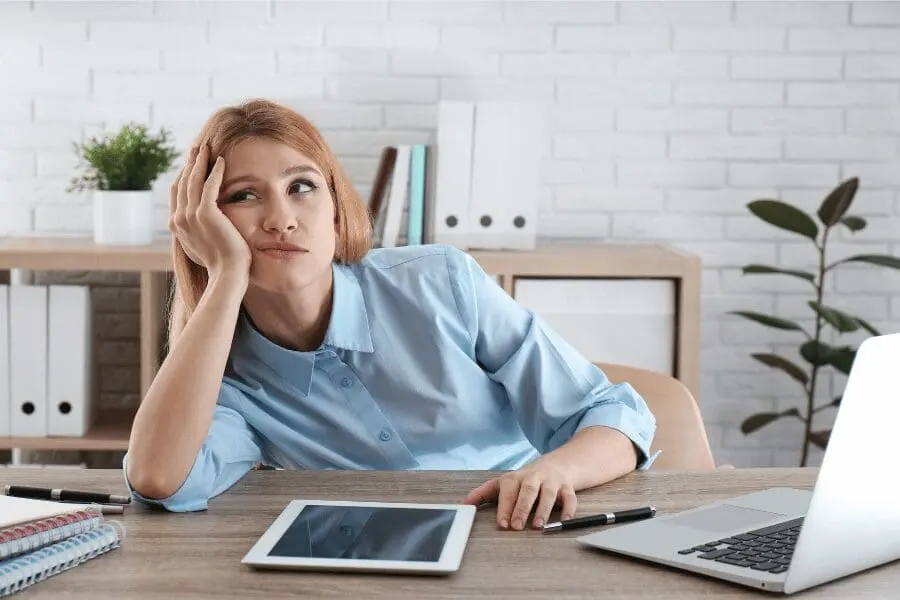 Une femme en chemise bleue est assise à un bureau, l'air ennuyé ou frustré, la tête appuyée sur sa main. Devant elle se trouvent une tablette, un cahier, un stylo et un ordinateur portable dans un bureau moderne.