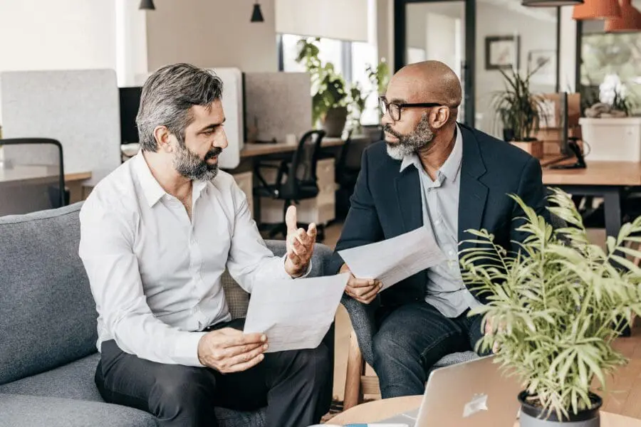 Deux hommes assis sur un canapé dans un bureau moderne, tenant des documents et ayant une discussion sérieuse, avec un ordinateur portable et une plante en pot sur la table devant eux.