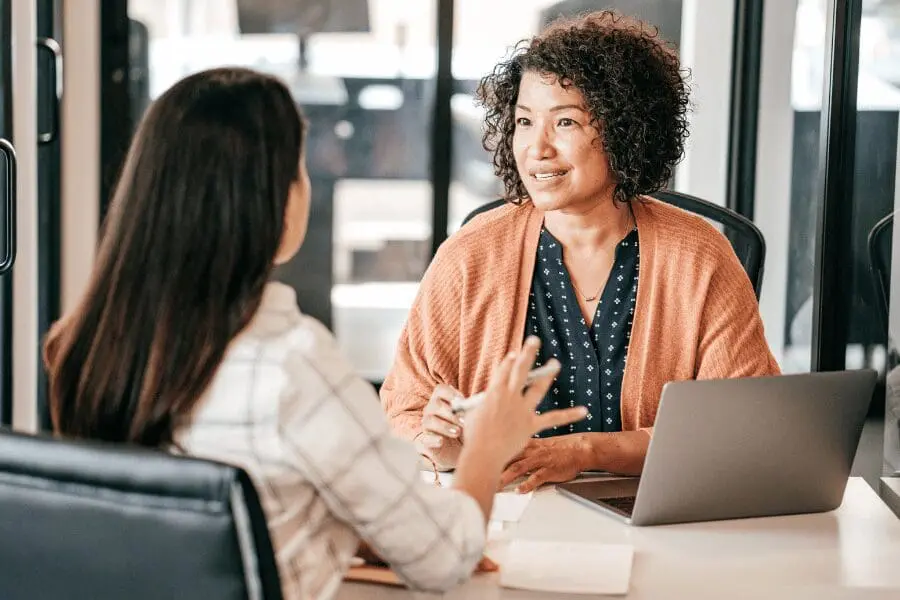 Deux femmes sont assises l'une en face de l'autre à un bureau et discutent. L'une d'elles a les cheveux bouclés et sourit tout en écoutant, son ordinateur portable ouvert devant elle.