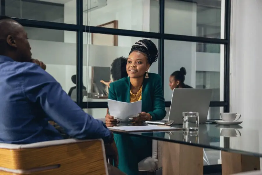 Une femme vêtue d'un blazer vert tient des documents et sourit tout en parlant avec un homme de l'autre côté d'une table en verre dans un bureau moderne. Un ordinateur portable, un verre d'eau et une tasse de café sont posés sur la table. Une autre personne travaille à l'arrière-plan.