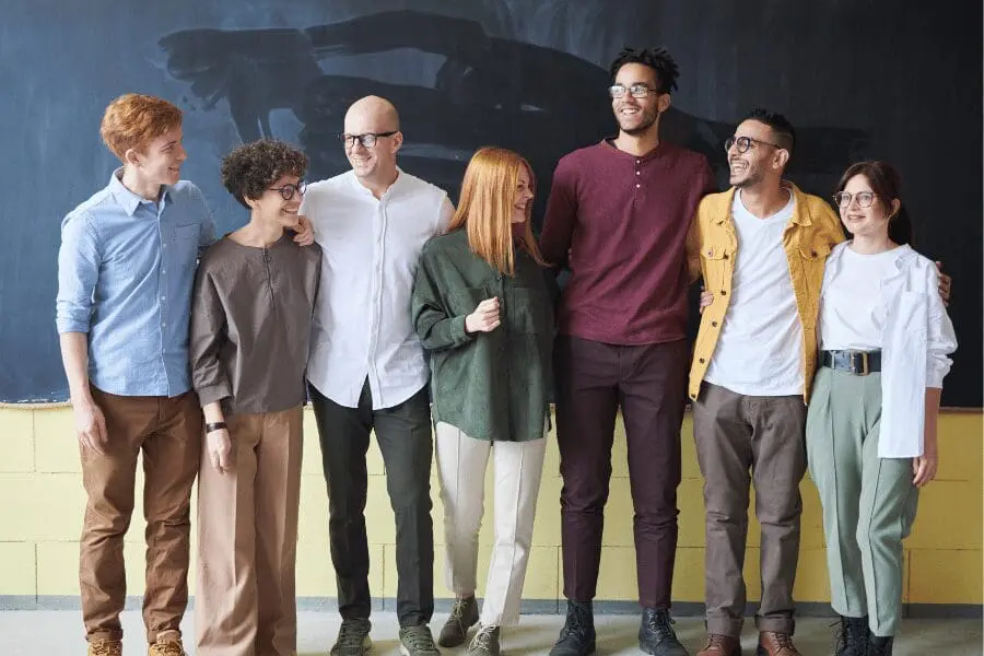 Six adultes se tiennent en rang, souriant et interagissant devant un tableau noir. Ils portent des vêtements décontractés dans des tons neutres et semblent amicaux et engagés les uns envers les autres.