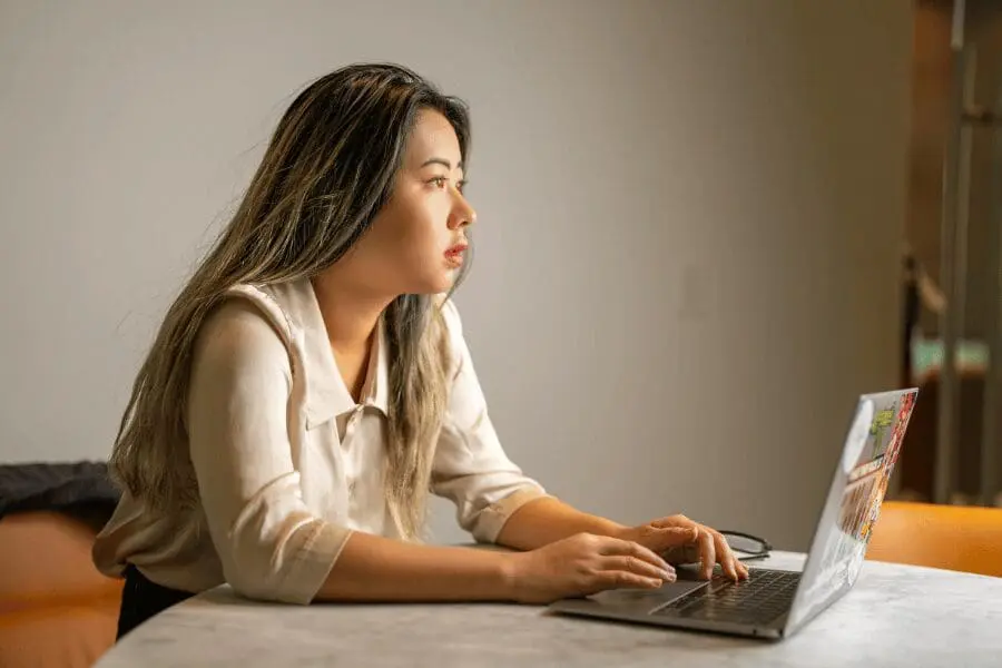 Une femme aux longs cheveux noirs est assise à une table et utilise un ordinateur portable. Elle regarde pensivement au loin, les mains posées sur le clavier. La scène se déroule à l'intérieur, avec un éclairage neutre.