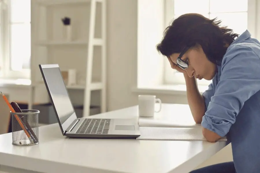 Une femme portant des lunettes est assise à un bureau avec un ordinateur portable et un carnet de notes ouverts, posant sa tête sur une main et semblant stressée ou fatiguée.