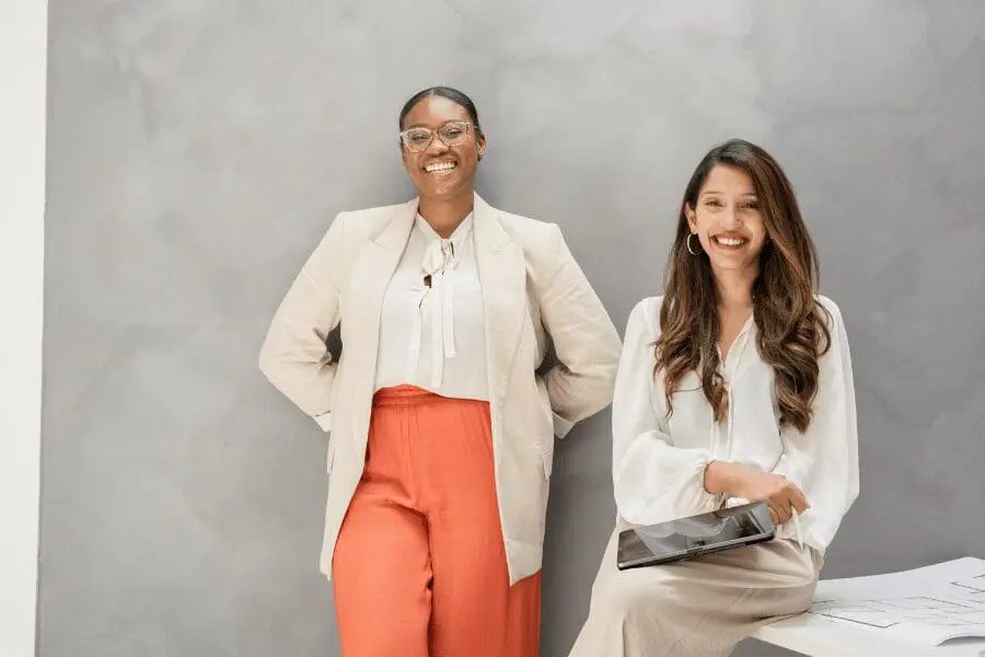 Deux femmes en tenue de travail sourient ; l'une est debout, les mains derrière le dos, vêtue d'un blazer beige et d'un pantalon orange, l'autre est assise, tenant une tablette, vêtue d'un chemisier blanc et d'une jupe beige, sur un fond gris clair.