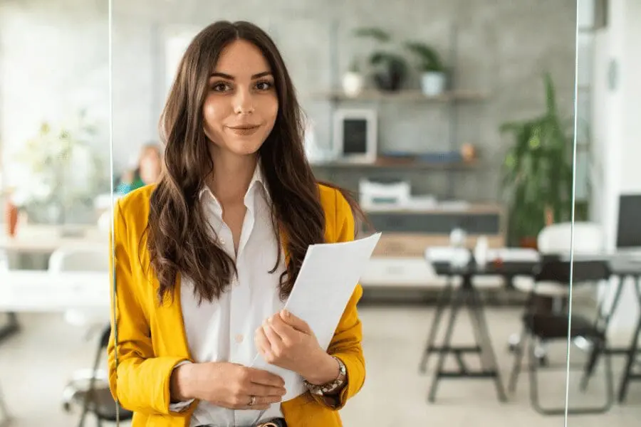 Une femme aux longs cheveux bruns, vêtue d'un blazer jaune et d'une chemise blanche, se tient dans un bureau et tient des documents. L'arrière-plan montre un bureau, des étagères et des plantes, créant ainsi un cadre moderne et professionnel.