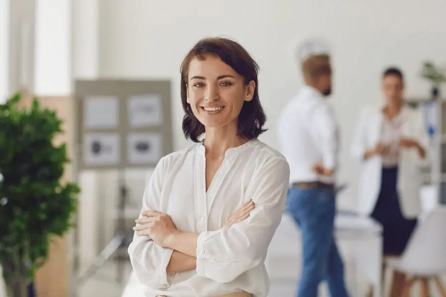 Une femme aux cheveux bruns courts et à la chemise blanche sourit, les bras croisés, dans un bureau. Dans l'arrière-plan flou, deux personnes discutent, il y a une plante et un tableau avec des papiers dessus.
