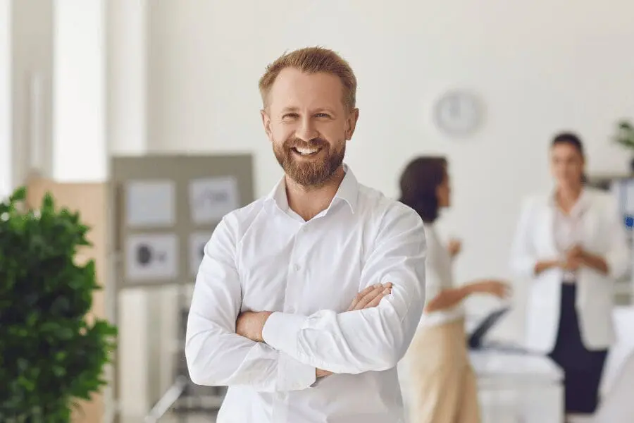 Un homme barbu et moustachu, vêtu d'une chemise blanche, sourit en croisant les bras dans un bureau. Deux personnes discutent dans l'arrière-plan flou, et il y a des plantes et une horloge dans la scène.