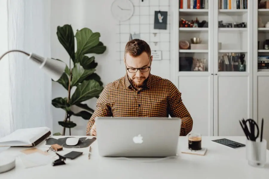 Un homme à lunettes est assis à un bureau blanc et travaille sur un ordinateur portable dans un bureau moderne et lumineux, avec des plantes, des étagères, des livres et des fournitures de bureau en arrière-plan.
