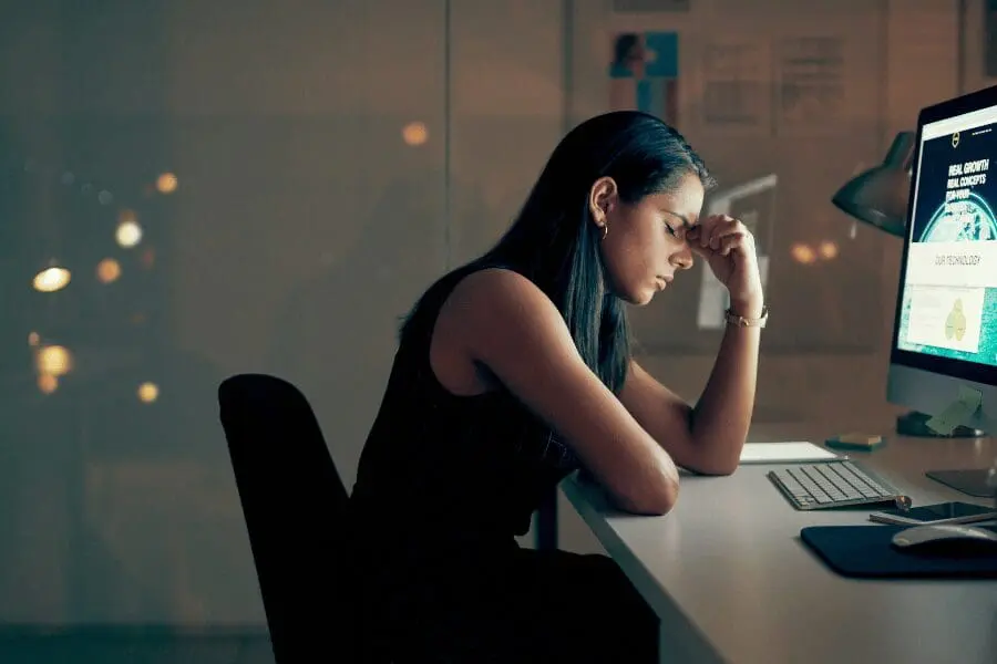 Dans un bureau faiblement éclairé, une femme est assise à un bureau, l'air stressé, la main sur le front, devant un écran d'ordinateur affichant un site web.
