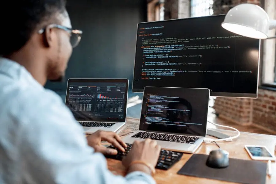 Une personne travaille à un bureau avec deux ordinateurs portables et un grand écran, tous affichant du code et des données. L'espace de travail comprend une lampe et une souris, et la personne tape sur un clavier.