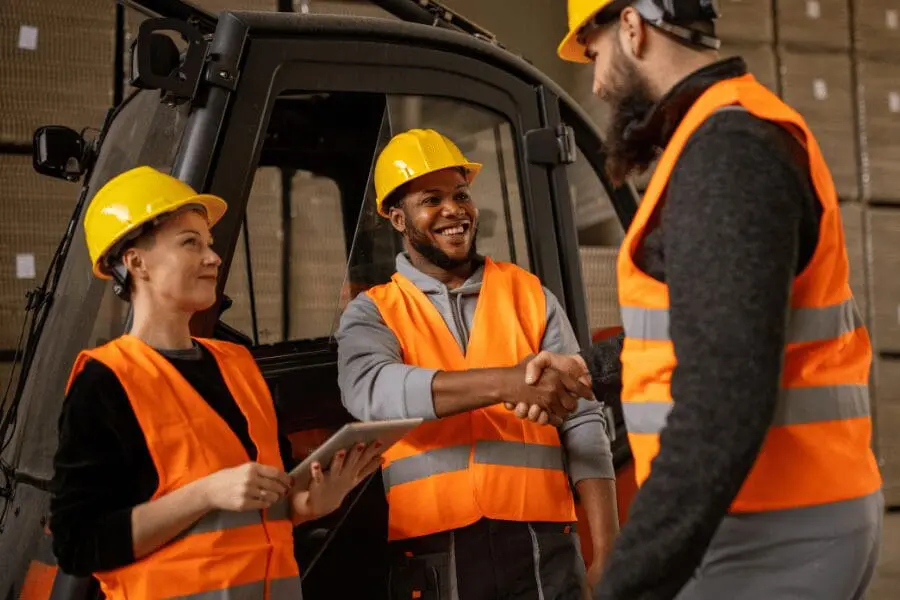 Trois ouvriers du bâtiment vêtus de gilets de sécurité orange et de casques jaunes sourient tandis que deux d'entre eux se serrent la main. L'un d'eux tient une tablette. Ils se tiennent devant des équipements industriels et des boîtes empilées.