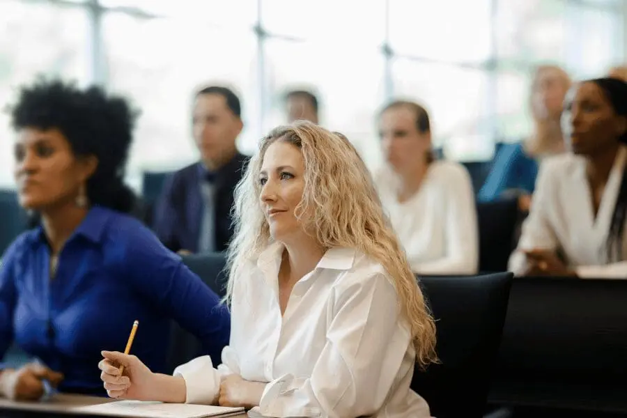 Une femme aux cheveux blonds bouclés, vêtue d'une chemise blanche, est assise à un bureau, tient un crayon et semble attentive dans une salle de classe, avec d'autres adultes à l'arrière-plan.