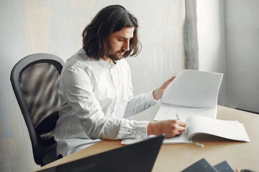 Un homme aux cheveux longs et à la chemise blanche est assis à un bureau, regardant des documents et écrivant des notes, avec un ordinateur portable et d'autres papiers à proximité.