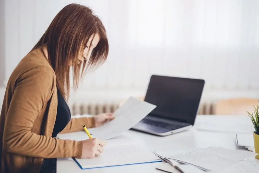 Une femme est assise à un bureau avec des papiers et un ordinateur portable, et écrit avec un stylo jaune. Elle semble concentrée, travaillant dans une pièce claire et bien éclairée, avec des rideaux blancs en arrière-plan.