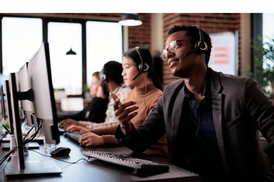 Trois agents d'un centre d'appel portant des écouteurs sont assis à des bureaux équipés d'ordinateurs, travaillant et discutant. Le cadre est celui d'un bureau moderne, éclairé par la lumière naturelle et de grandes fenêtres en arrière-plan.