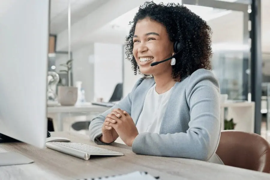 Une femme souriante portant un casque est assise à un bureau et regarde un écran d'ordinateur, ce qui suggère qu'elle est en train de participer à un appel vidéo ou à une réunion virtuelle.