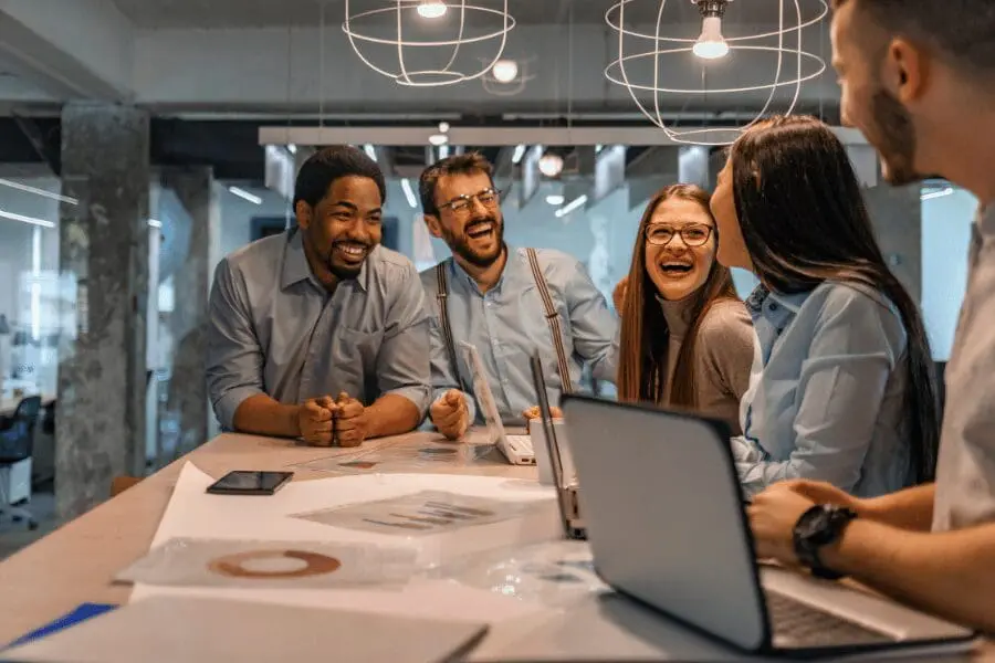 Cinq collègues, habillés de manière décontractée, riant et discutant autour d'une table avec des ordinateurs portables, des documents et des tableaux dans un bureau moderne avec des lumières suspendues au-dessus d'eux.