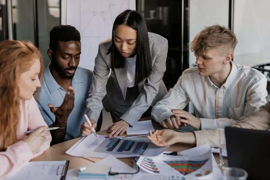 Quatre personnes sont assises à une table couverte de tableaux et de graphiques, tandis qu'une femme debout se penche pour discuter et montrer un document du doigt. Ils semblent collaborer à un projet d'entreprise.