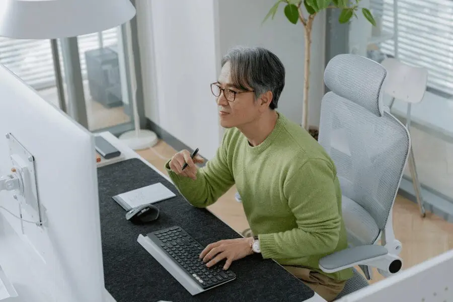 Un homme portant des lunettes et un pull vert est assis à un bureau, travaillant sur un ordinateur avec un clavier et une souris. Des notes et un stylo sont posés sur le bureau, et une plante est visible à l'arrière-plan.