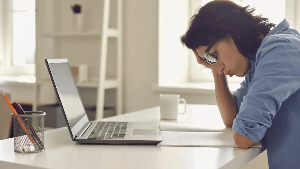 Une femme portant des lunettes est assise à un bureau, la tête appuyée sur sa main, l'air fatigué ou stressé. Un ordinateur portable ouvert, un carnet de notes et une tasse se trouvent devant elle dans une pièce lumineuse et ensoleillée.