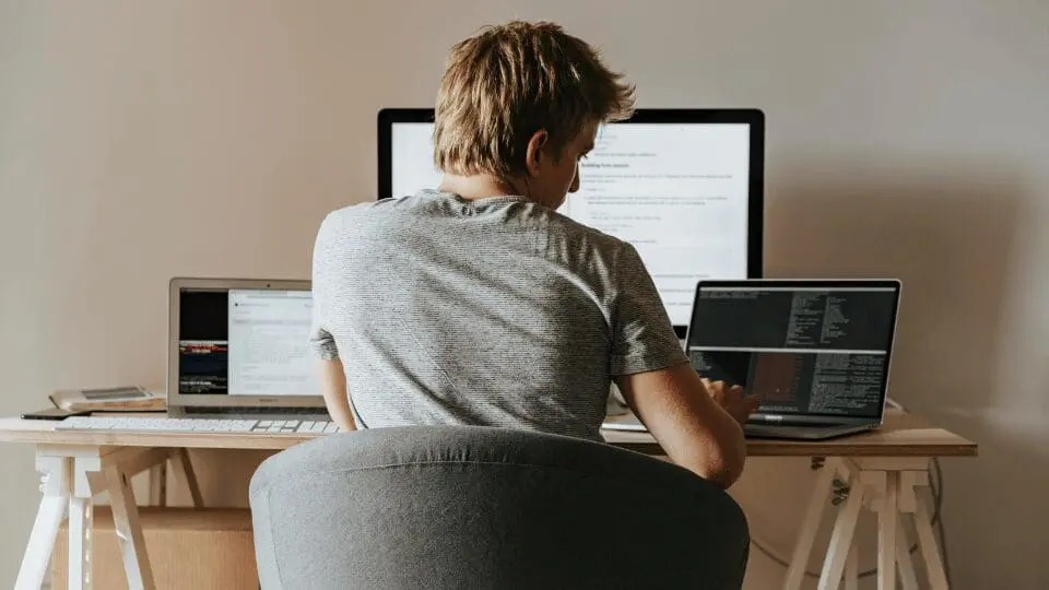 Une personne aux cheveux courts et clairs est assise à un bureau tourné vers l'extérieur et travaille sur un ordinateur portable dont l'écran affiche un code. Deux autres écrans et un clavier se trouvent également sur le bureau dans une pièce moderne et bien éclairée.
