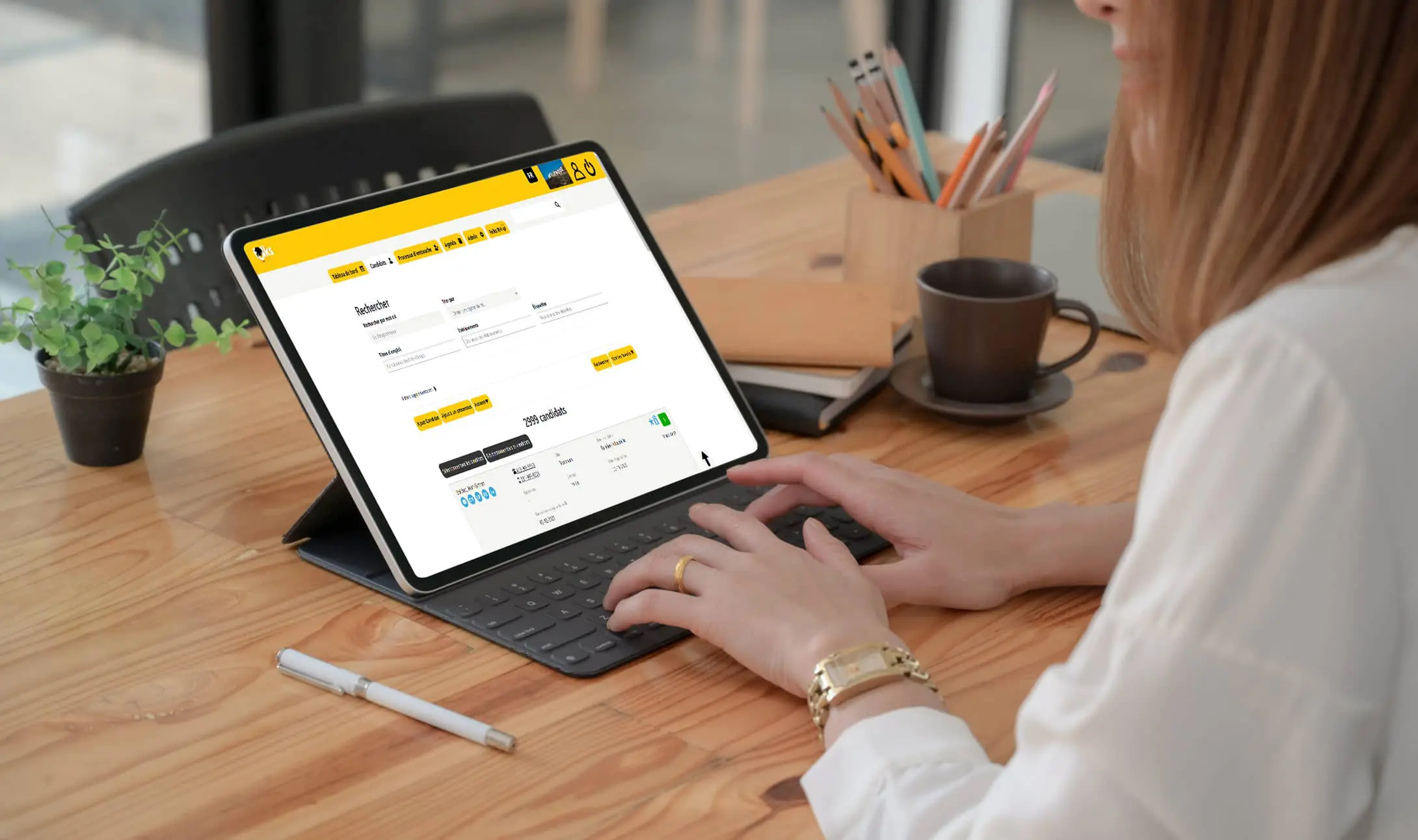 Une femme tape sur une tablette avec clavier, assise sur un bureau en bois, et consulte un site de banque en ligne. À proximité se trouvent une tasse de café, un carnet, un stylo et une paire de lunettes.