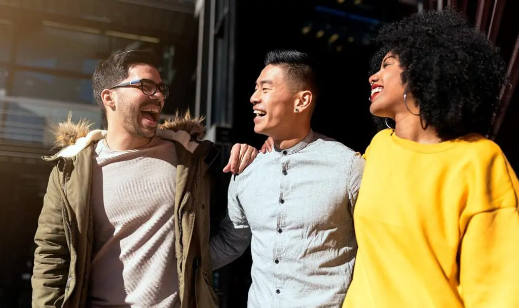 Trois amis se promènent dehors au soleil, souriant et riant ensemble. L'homme à gauche porte des lunettes et un manteau, celui du milieu une chemise claire et la femme à droite un pull jaune vif.
