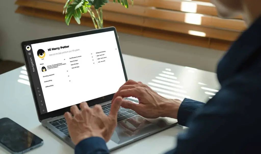 A person wearing a dark shirt types on a laptop at a white desk. The laptop screen displays a profile page with the name