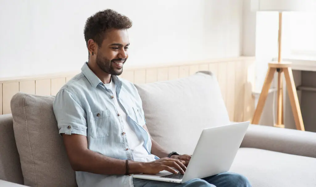A smiling man sits on a beige sofa using a laptop, wearing a light denim shirt and blue jeans. Natural light enters from a window beside him, and a tripod lamp stands in the background.
