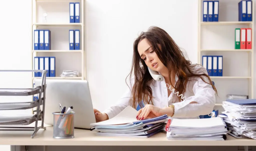 Une femme est assise à un bureau encombré, parle au téléphone, tape sur un ordinateur portable et trie des documents, semblant stressée et occupée dans un bureau avec des étagères de classeurs en arrière-plan.