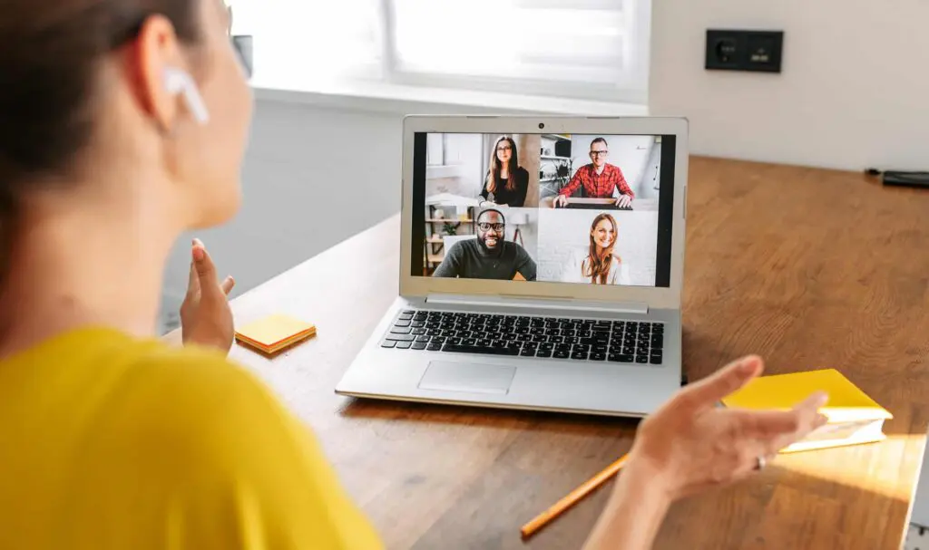 A woman wearing earbuds sits at a desk, participating in a video call on her laptop with four people visible on the screen. Sunlight comes through a window in the background.