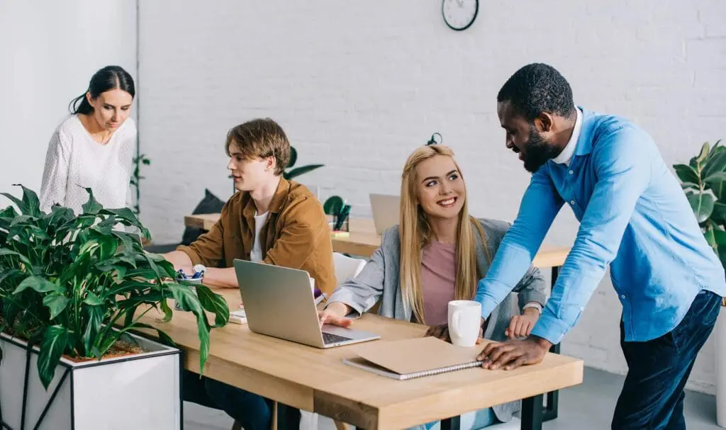 Quatre personnes travaillent ensemble dans un bureau moderne avec des bureaux en bois et des plantes. Deux personnes sont assises à un bureau avec un ordinateur portable, une autre discute debout, et une autre travaille en arrière-plan. L'atmosphère est collaborative et conviviale.