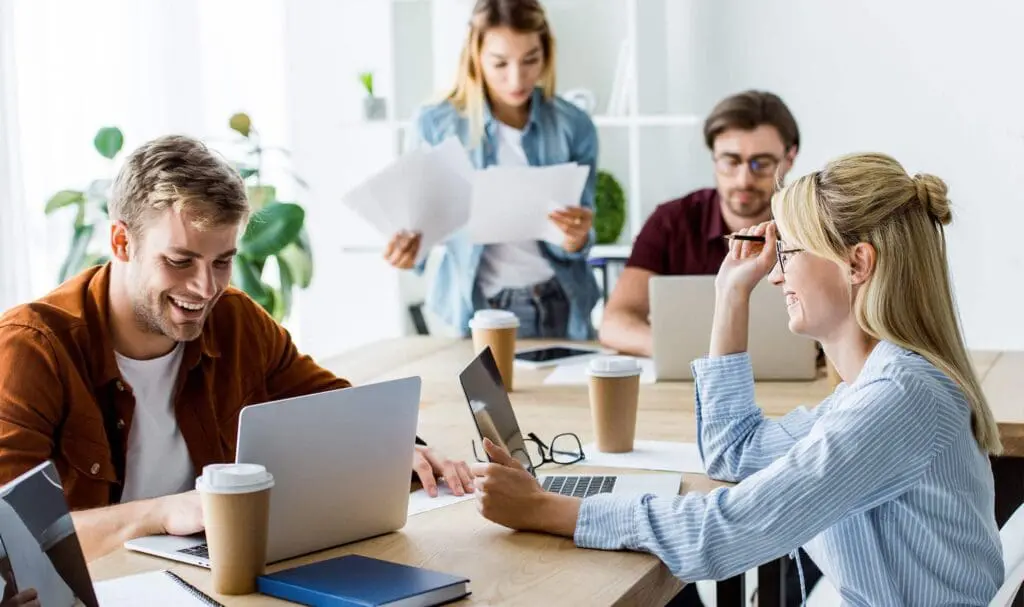 Quatre jeunes adultes travaillent ensemble à une table avec des ordinateurs portables, des papiers et des tasses à café. Deux sont assis et sourient, un est debout en train de lire des documents, et un autre travaille sur un ordinateur portable dans un bureau lumineux.
