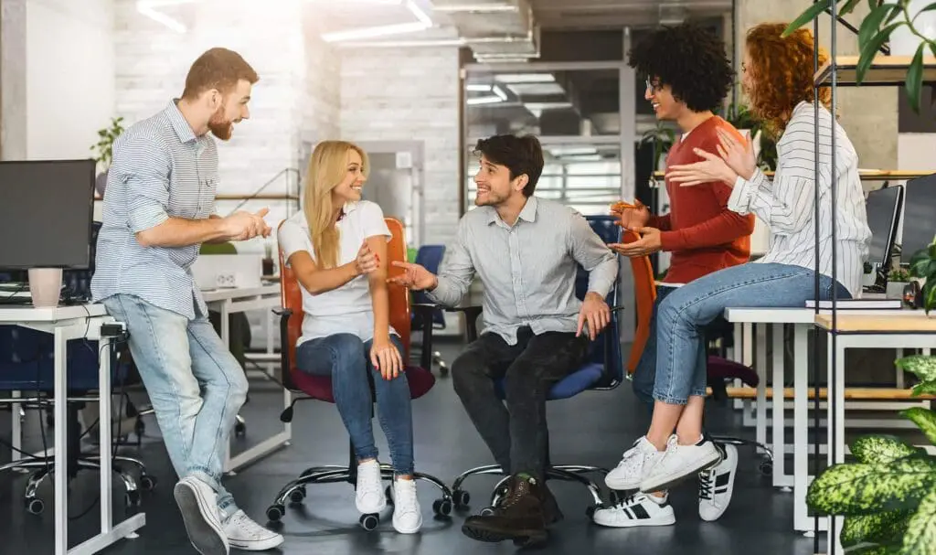 Cinq jeunes adultes en tenue décontractée sourient et discutent ensemble dans un espace de bureau moderne et lumineux, assis sur des chaises et des bureaux, créant une atmosphère conviviale et collaborative.