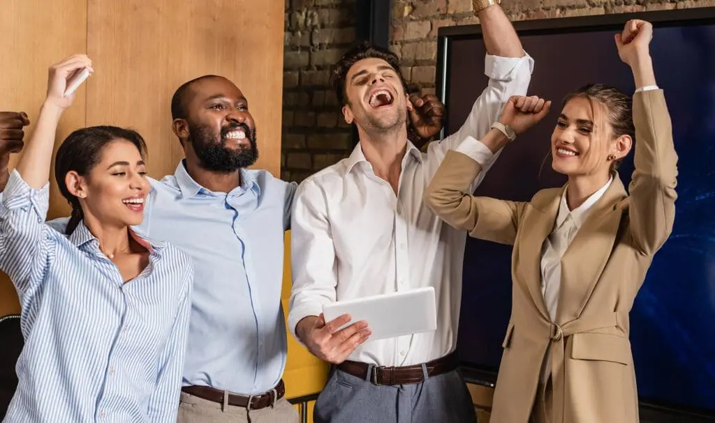 Quatre personnes, habillées de façon professionnelle, se tiennent côte à côte à l'intérieur, souriantes et levant les bras en signe de célébration. L'une d'elles tient une tablette. Elles paraissent heureuses et enthousiastes, suggérant une réussite ou une victoire d'équipe.