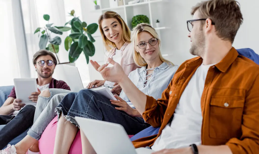 Quatre jeunes adultes assis sur un canapé avec ordinateurs portables et tablettes, souriant et discutant dans une pièce lumineuse et fleurie. Ils semblent engagés et collaboratifs.