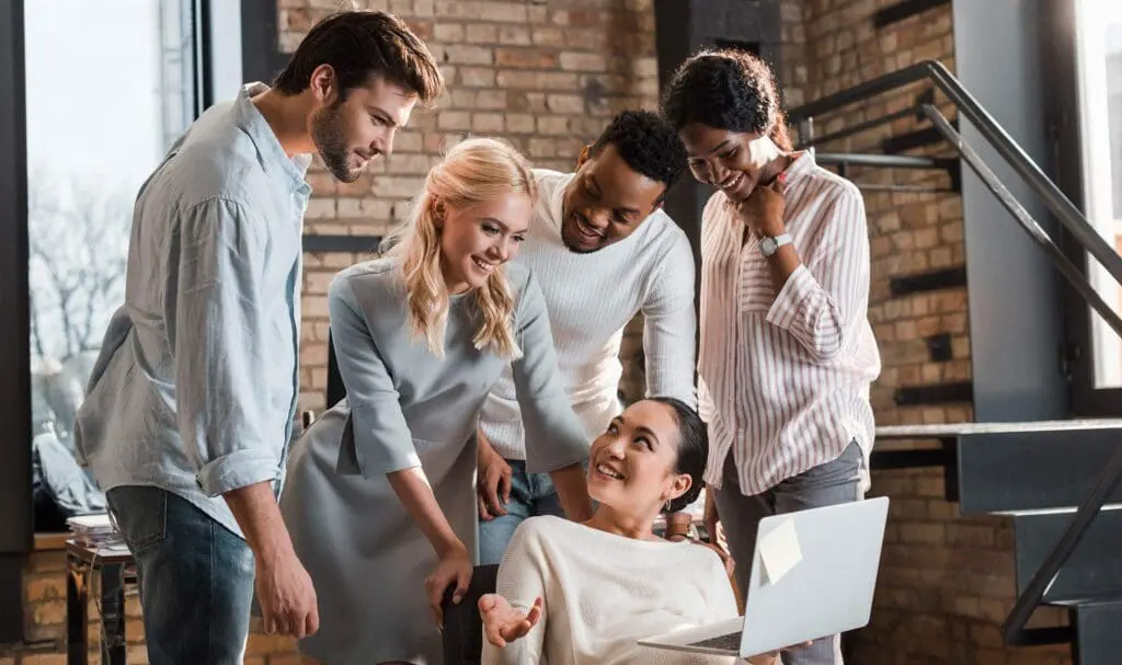 Cinq personnes se rassemblent autour d'un ordinateur portable, souriantes et engagées dans une conversation, dans un bureau moderne avec des murs en briques et de grandes fenêtres.