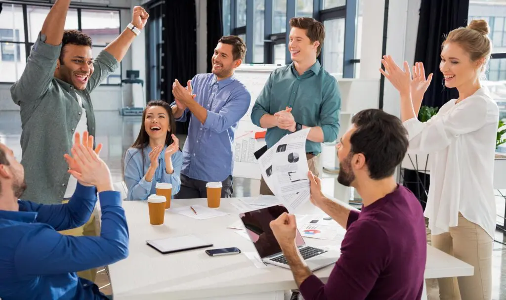 A group of six colleagues sit and stand around a table in a bright office, smiling and clapping in celebration. Laptops, papers, and coffee cups are on the table, suggesting a successful meeting.