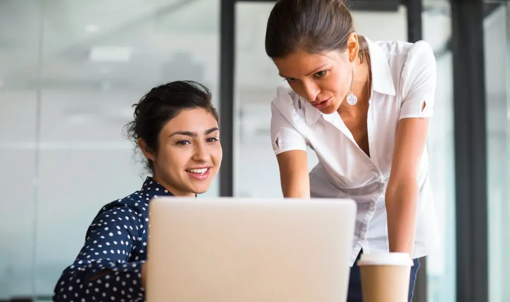 Two women work together at a desk with a laptop. One woman is sitting and smiling at the screen, while the other stands beside her, pointing at the laptop. Coffee cups are also visible on the desk.
