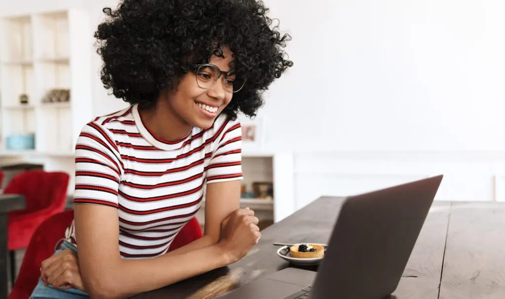 A smiling woman with curly hair and glasses, wearing a striped shirt, sits at a table looking at a laptop. A small dessert is on a plate nearby. The setting is bright and modern.