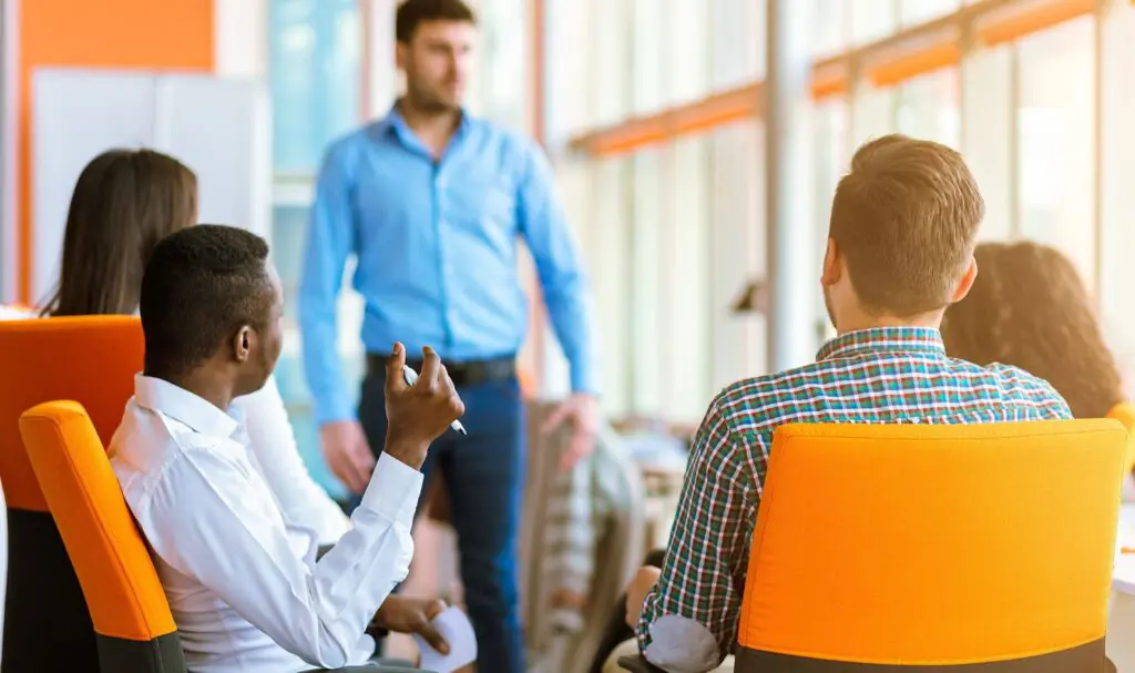 Un groupe de personnes assises sur des chaises orange écoutent un homme debout et parlant dans un bureau lumineux et moderne avec de grandes fenêtres.