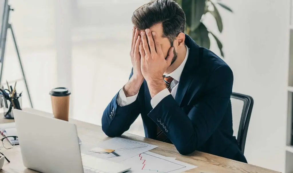 Un homme en costume est assis à un bureau, les mains sur le visage, l'air stressé. Un ordinateur portable ouvert, des documents avec des graphiques, un stylo, un carnet et une tasse de café à emporter sont posés sur le bureau.