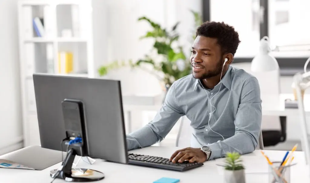 A man wearing earphones smiles while working at a desktop computer in a bright, modern office. There are plants, office supplies, and shelves in the background.