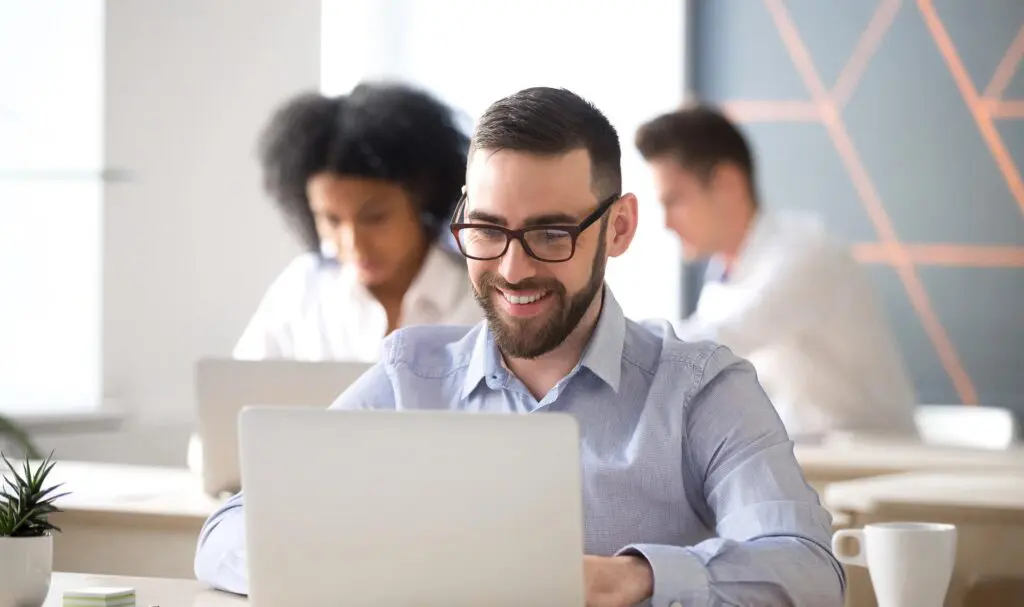 Un homme portant des lunettes et une barbe sourit en travaillant sur un ordinateur portable à son bureau. Deux autres personnes travaillent en arrière-plan. Le cadre est lumineux et moderne.