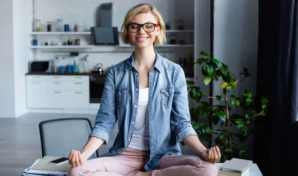 A smiling woman with glasses sits cross-legged on a desk in a modern kitchen, meditating with her eyes closed and hands resting on her knees, surrounded by books and a potted plant.